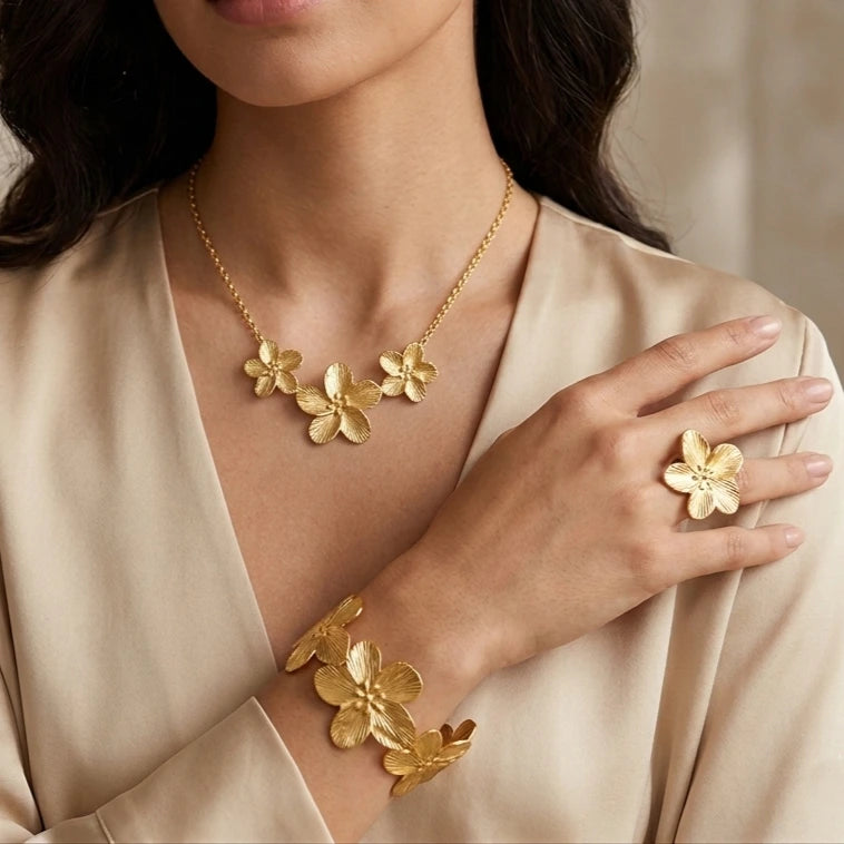 Portrait rapproché d'une femme portant la parure complète dorée Colette - Épanouie. Le collier à trois fleurs, la bague imposante et le bracelet manchette assorti présentent des pétales finement ciselés qui capturent la lumière sur une tenue en satin.