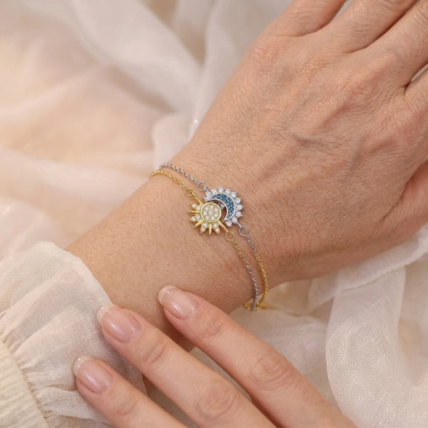 Gros plan sur un poignet féminin portant le duo de bracelets Hélios (soleil doré) et Séléné (lune bleue argentée) superposés sur un tissu en mousseline blanche.